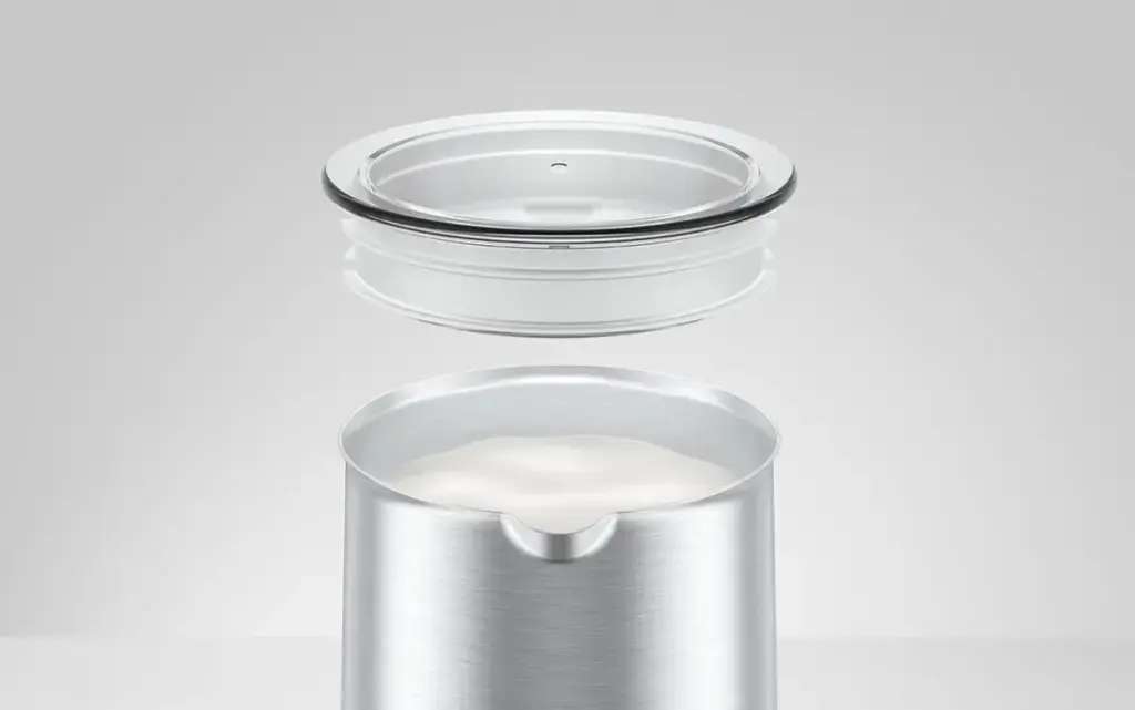 מקציף חלב אוטומטי של JURA (חם וקר) עשוי מתכת ממוקם מתחת למכסה עגול ושקוף המרחף מעליו, על רקע אפור בהיר ופשוט. | אספרסימו