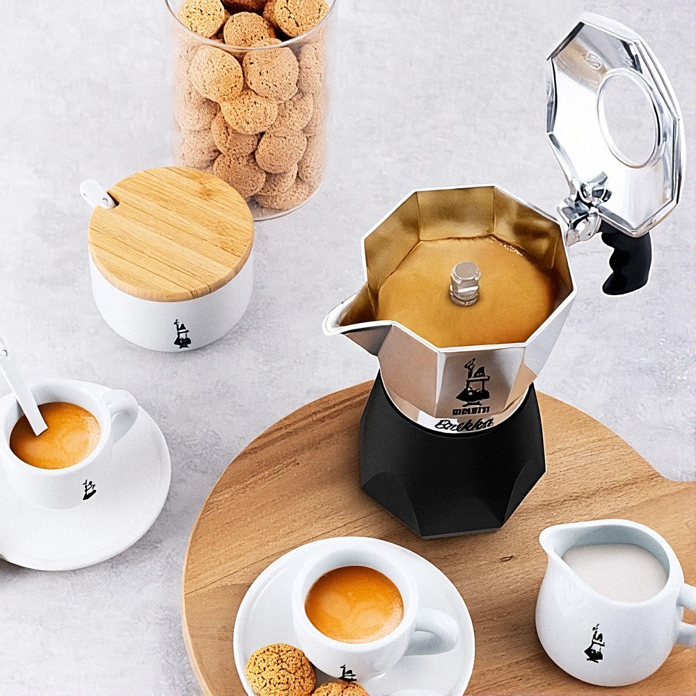 מקינטה מסוג "ביאלטי בריקה" (Bialetti Brikka) ל-2 כוסות, מלאה בקפה, מונחת על השולחן לצד שתי כוסות אספרסו, קנקן חלב, צנצנת עוגיות וקערת סוכר — כולם מתאפיינים בגוונים תואמים של לבן ועץ, היוצרים אווירה מזמינה. | אספרסימו