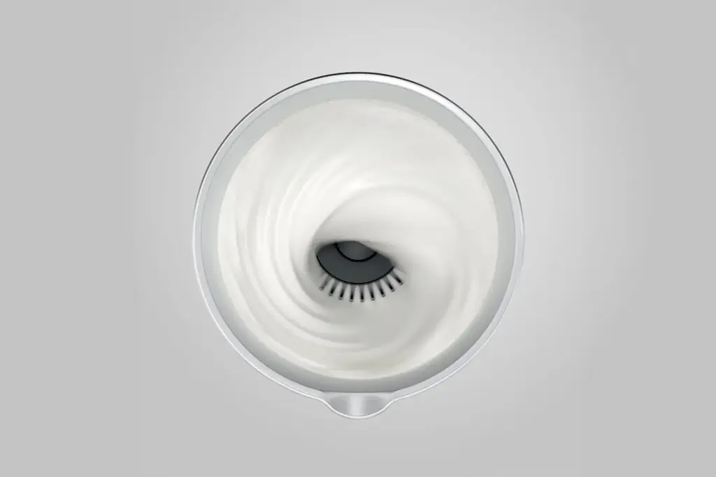 תצוגה מלמעלה של מקציף החלב האוטומטי – חם וקר של JURA בפעולה, היוצר מערבולת מסחררת של חלב לבן וקצפי בתוך מיכל המתכת העגול שלו, על רקע אפור בהיר. | אספרסימו